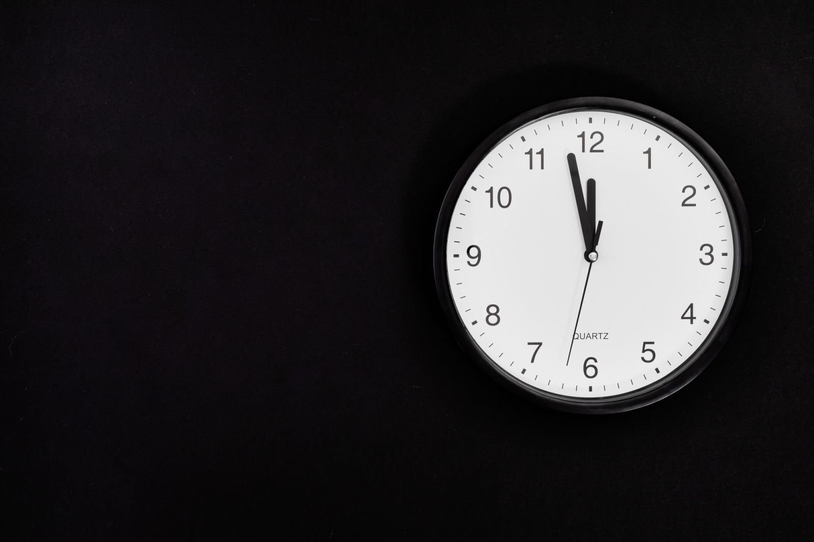 Black-framed wall clock reading just before midnight against a black background