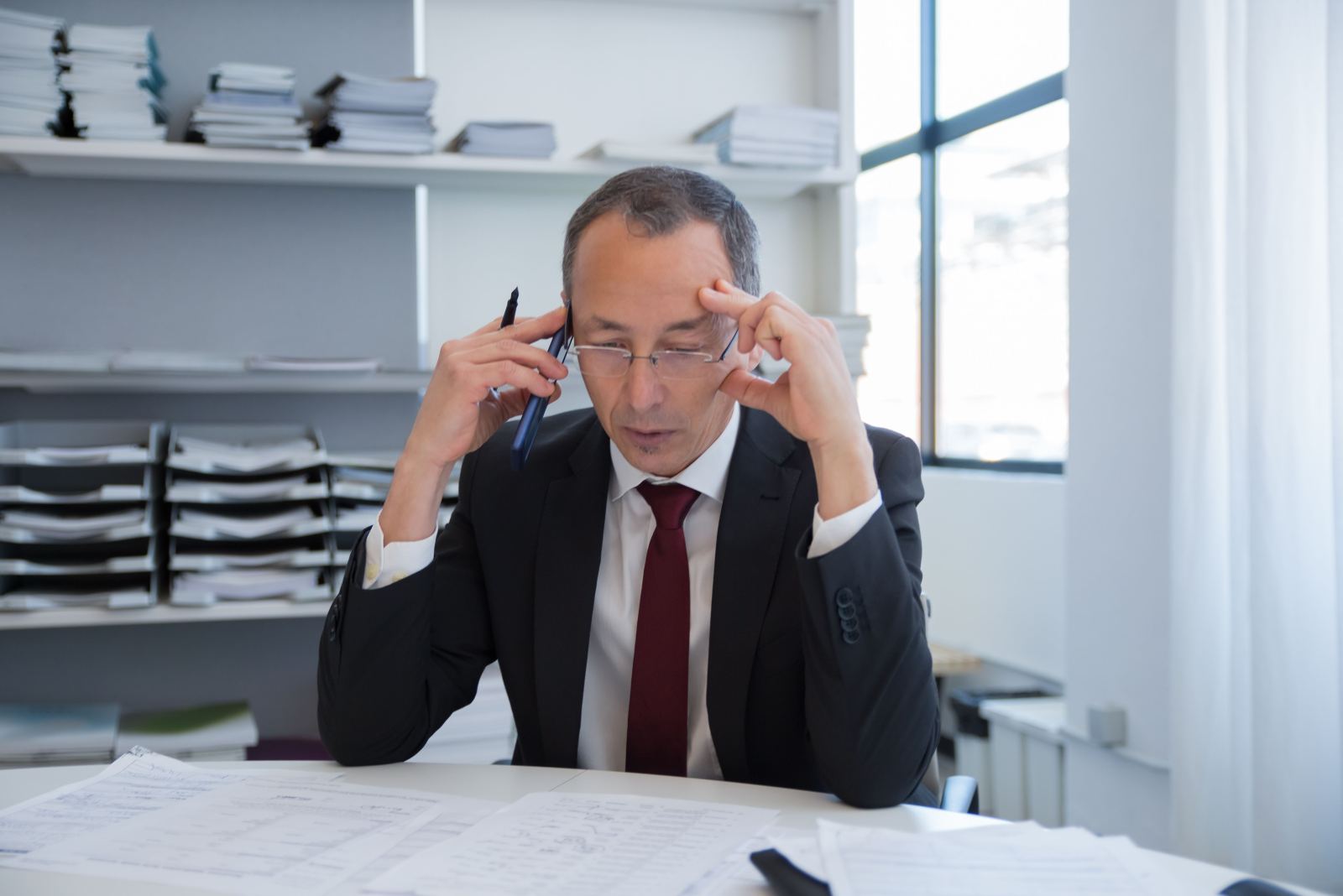 Businessman rubbing his forehead while on a phone call surrounded by paperwork