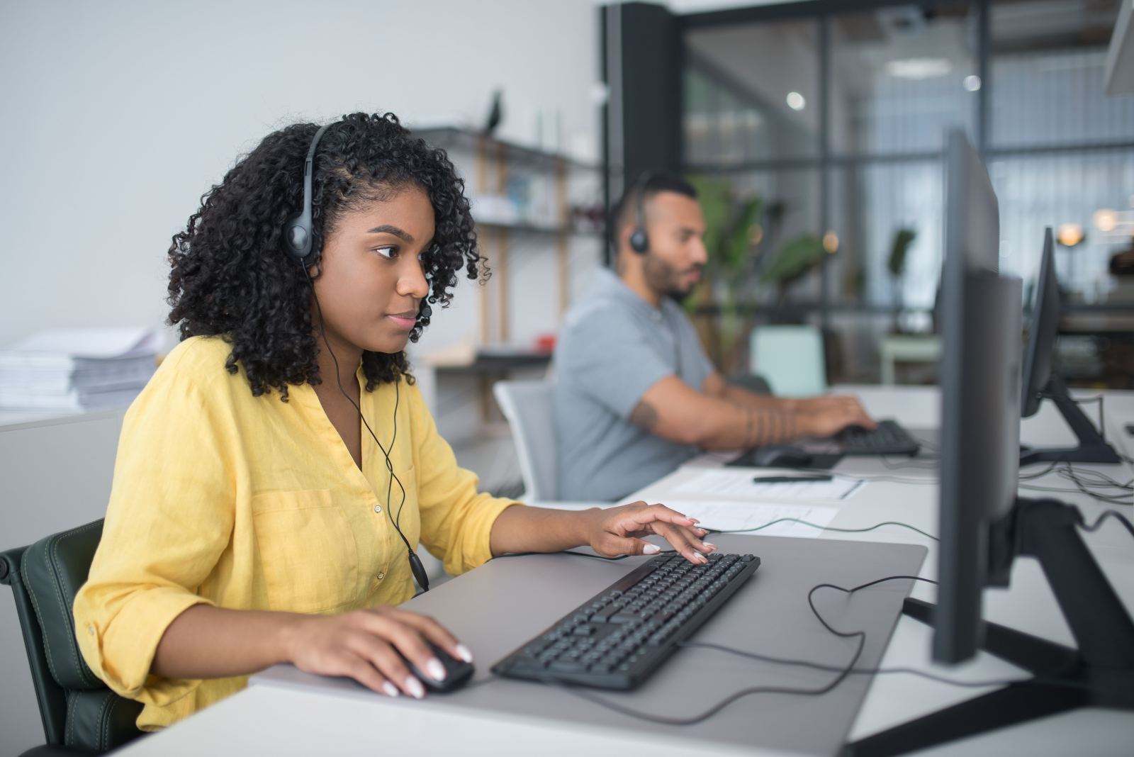 Call center agent in a yellow shirt working at a computer with a colleague visible behind her