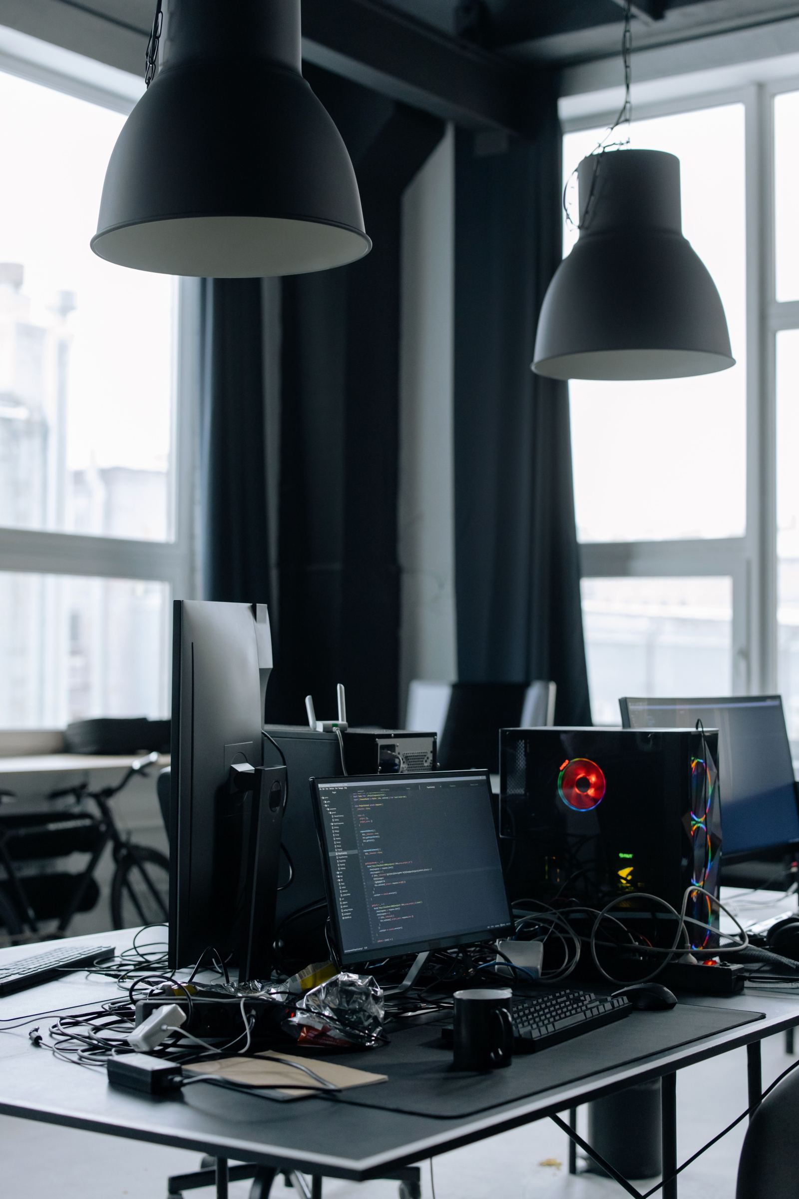 Office desk with a code editor open on a monitor beneath hanging pendant lamps