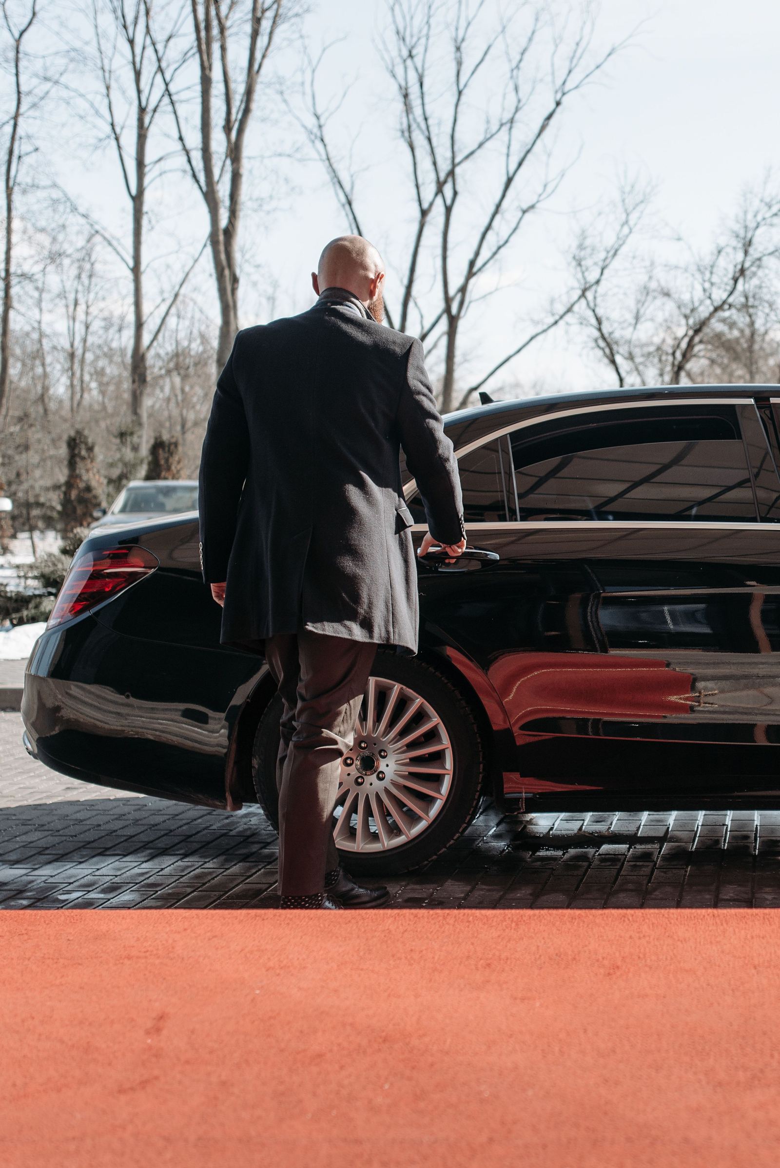 Chauffeur in a coat approaching a black luxury car on a red carpet