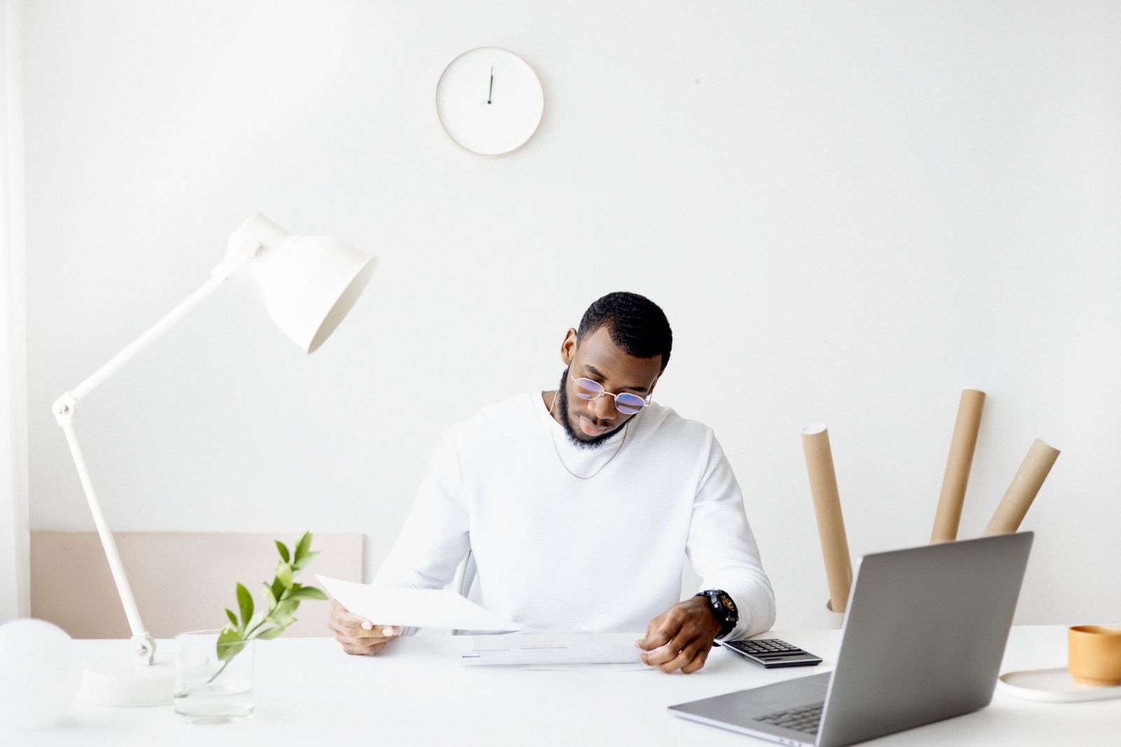 Man reviewing papers at a desk beneath a wall clock in a bright office