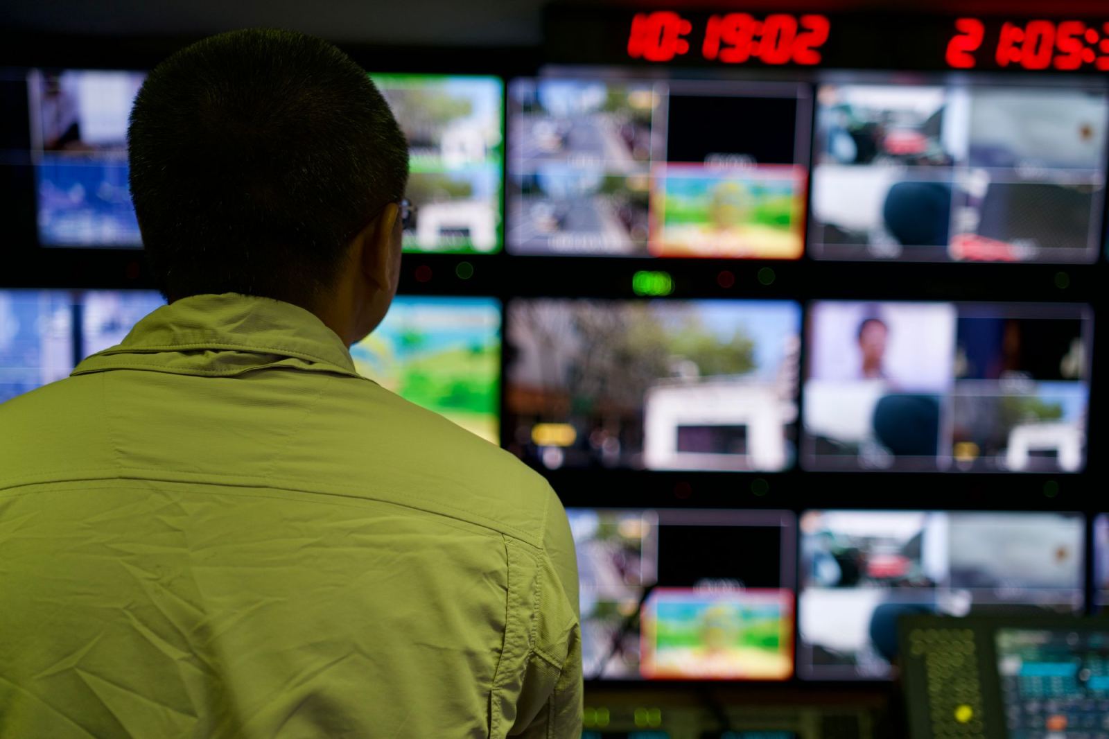Man viewed from behind watching a wall of monitors in a control room
