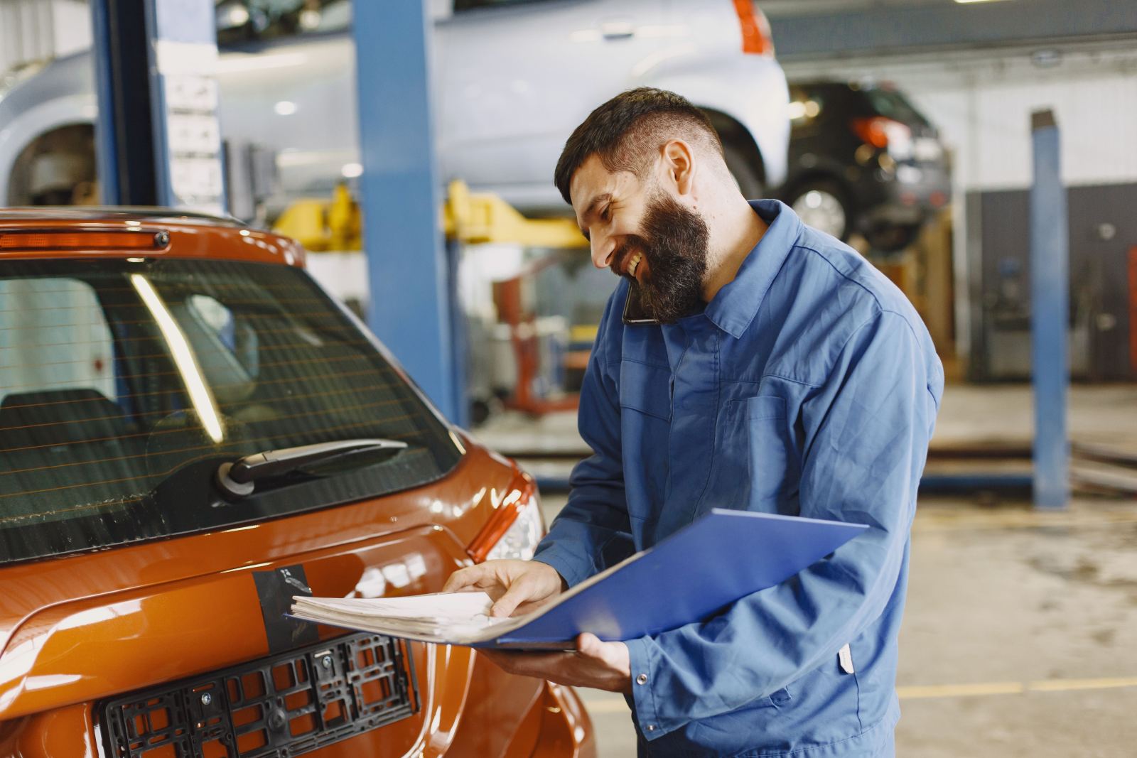 Mechanic smiling while on a phone call and holding a clipboard in a garage