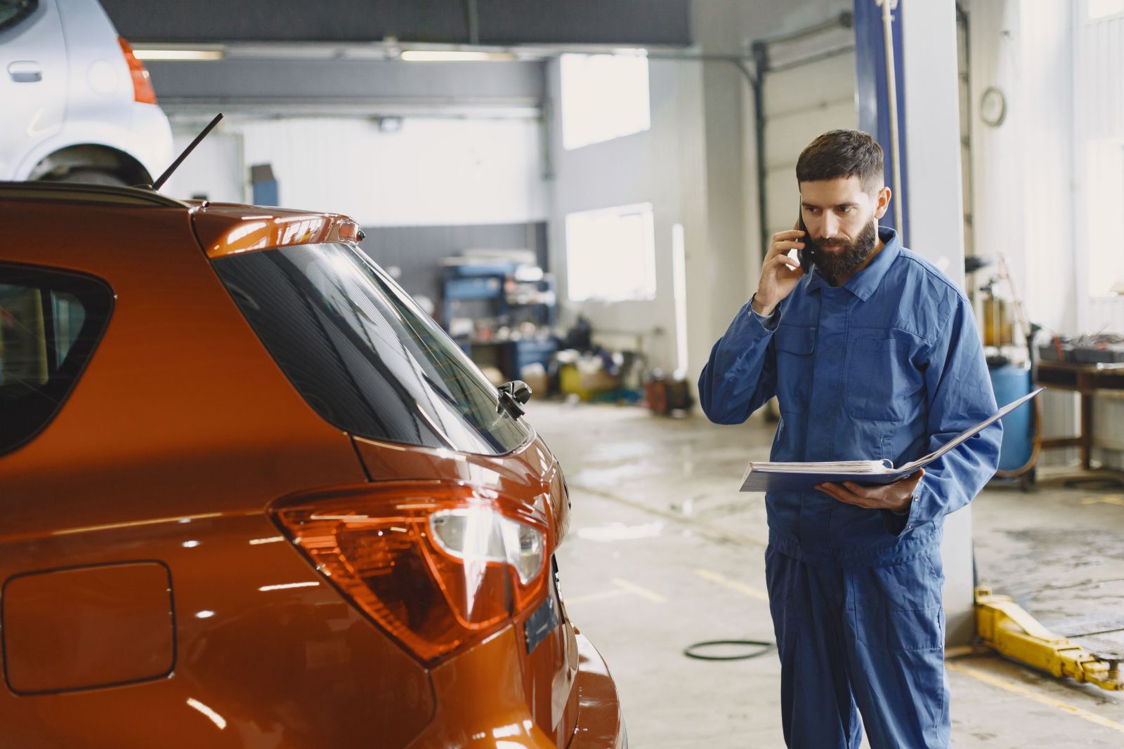 Mechanic on a phone call holding a clipboard beside an orange car in a garage