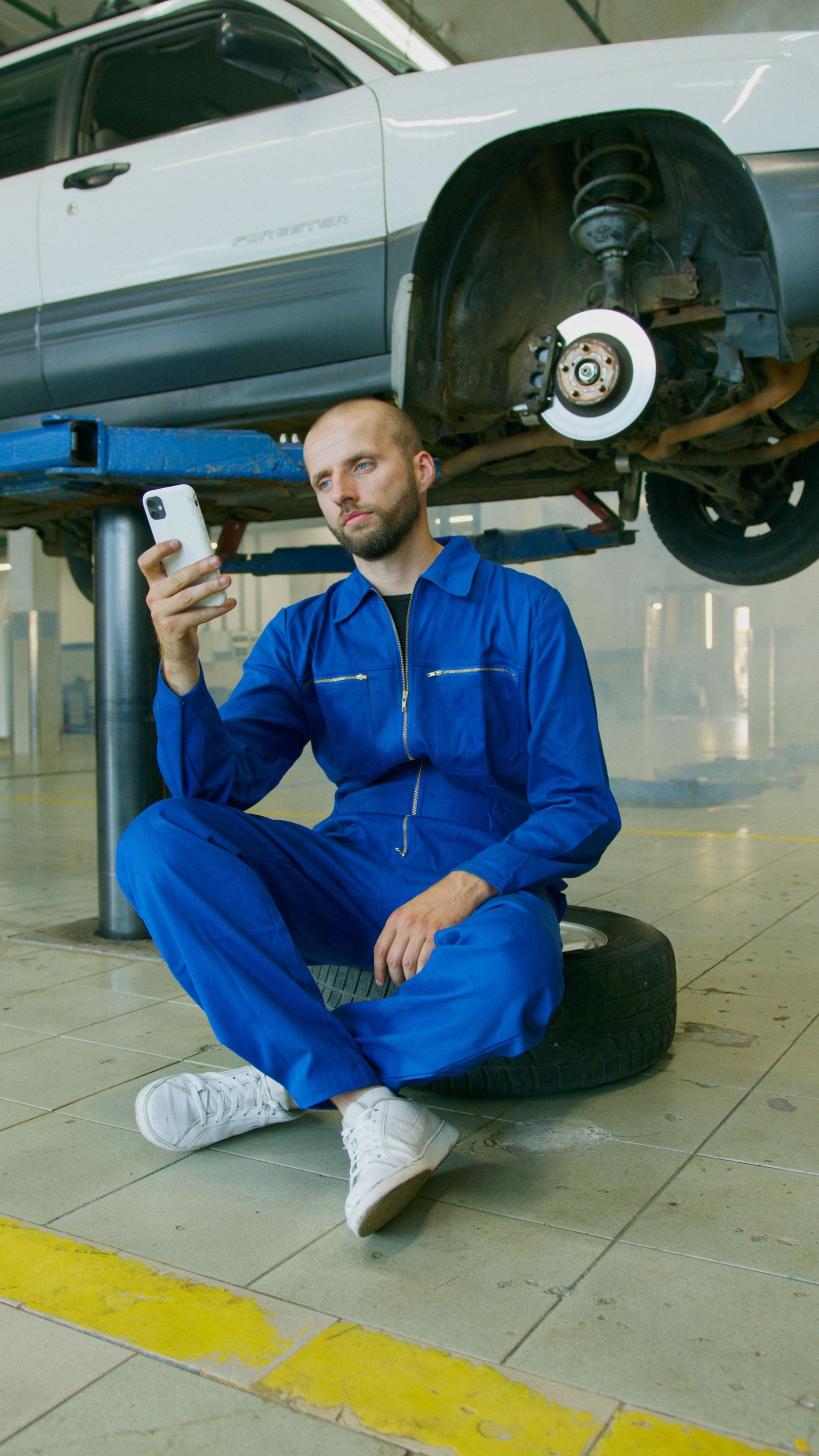 Mechanic sitting on a tire checking his phone beneath a car raised on a lift