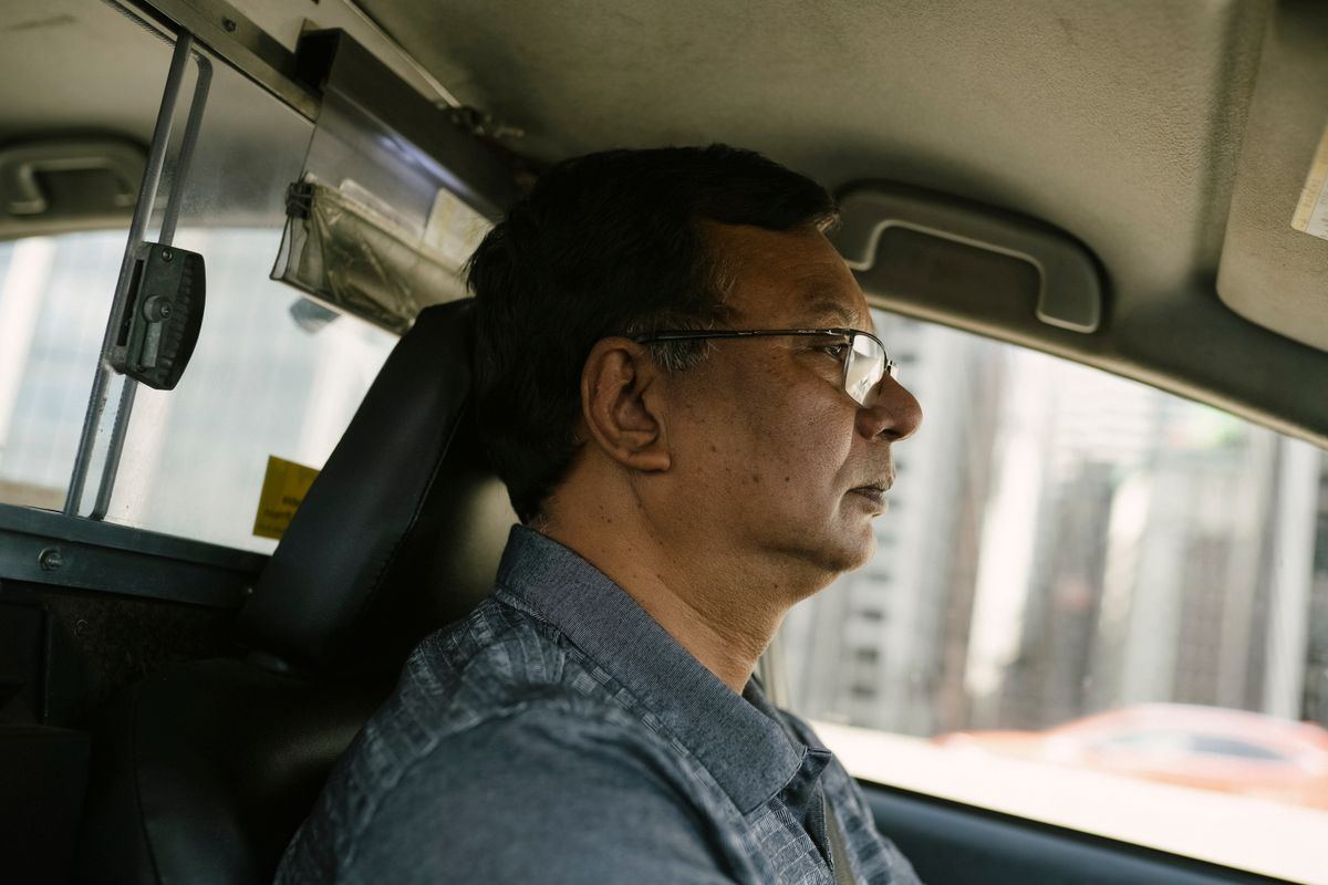 Taxi driver wearing glasses, focused, city skyscrapers visible through the window