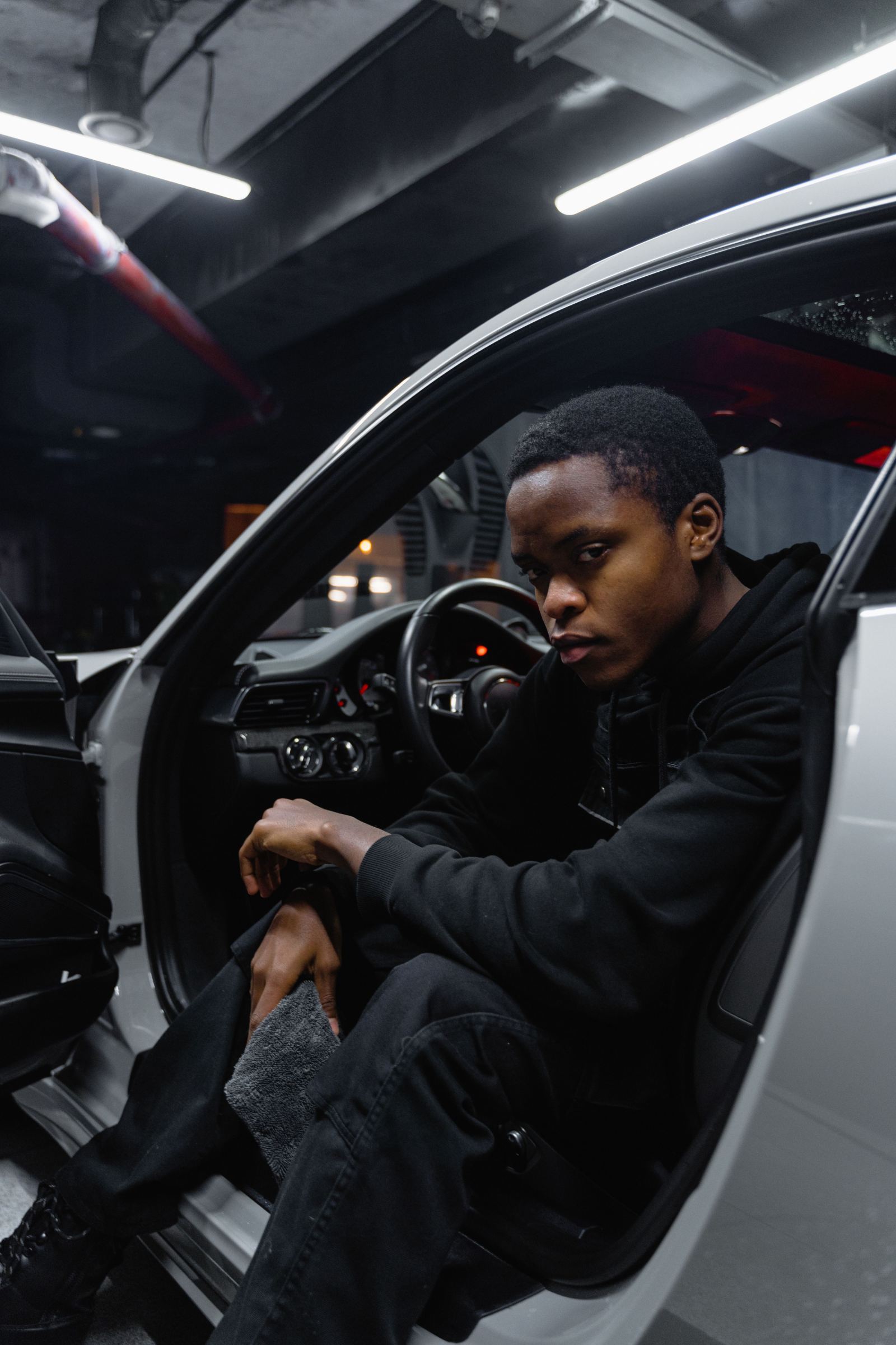 Man in a dark hoodie sitting in a car with the door open in a garage