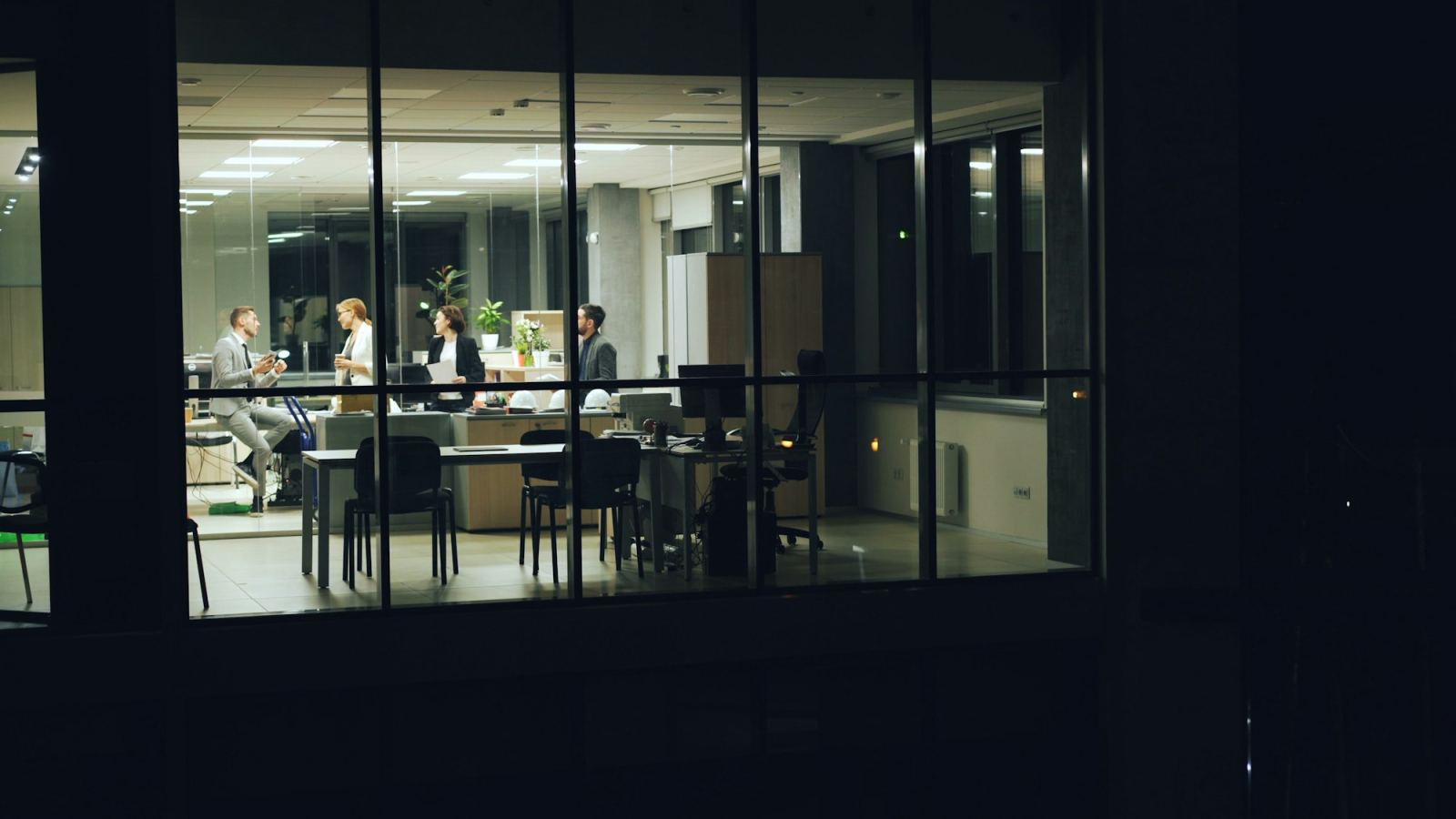 Team of colleagues talking in a lit office at night, seen through a large window