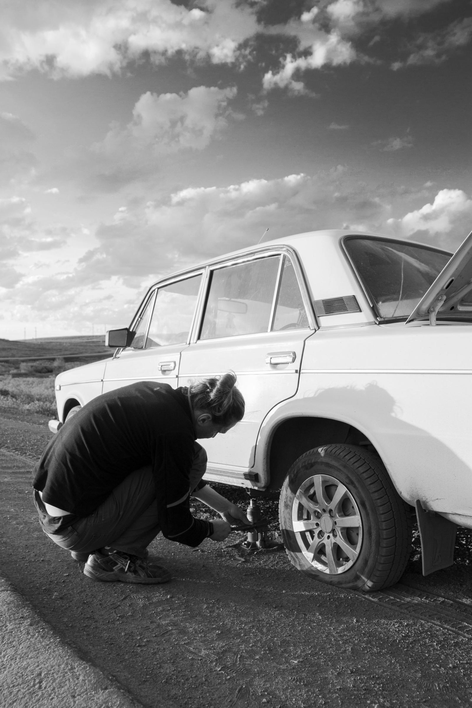 Black-and-white photo of a driver changing a tire on a rural road