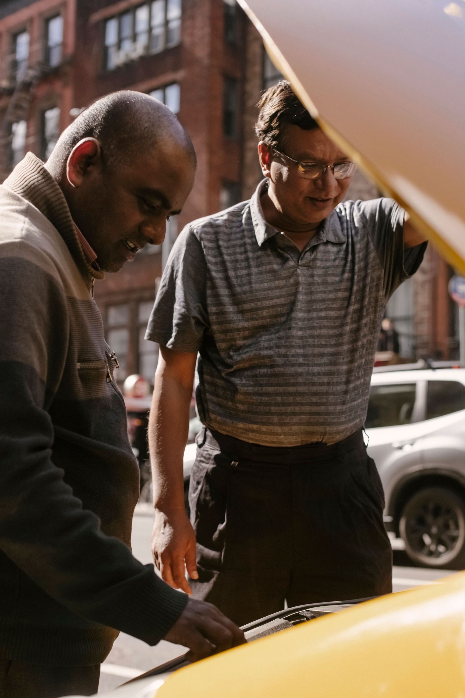 Two men looking under the hood of a yellow taxi on a city street