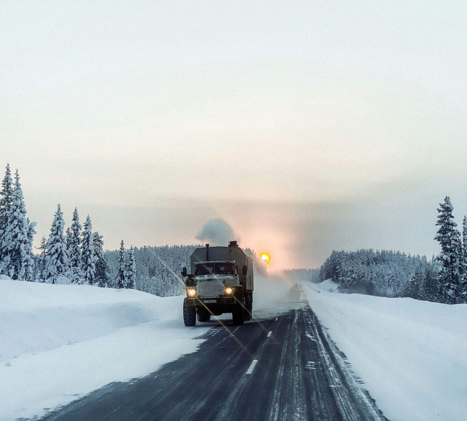 Truck driving down a snowy road with exhaust visible at sunset