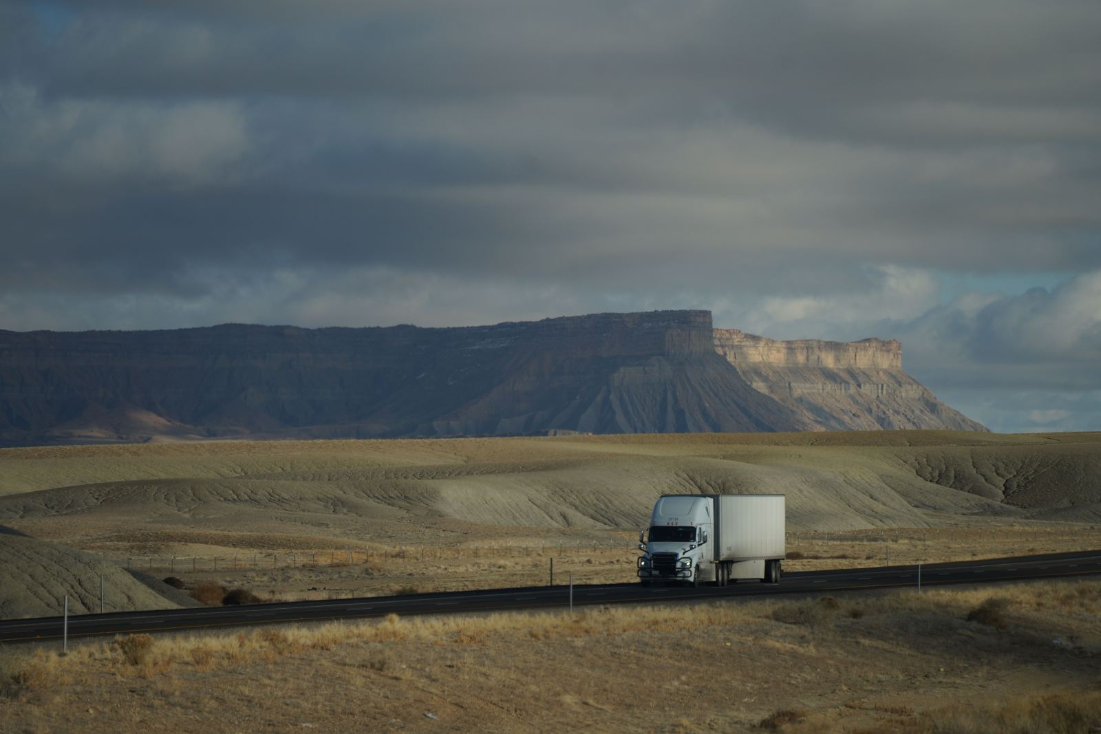A single semi truck driving alone on a desert highway beneath dramatic cliffs