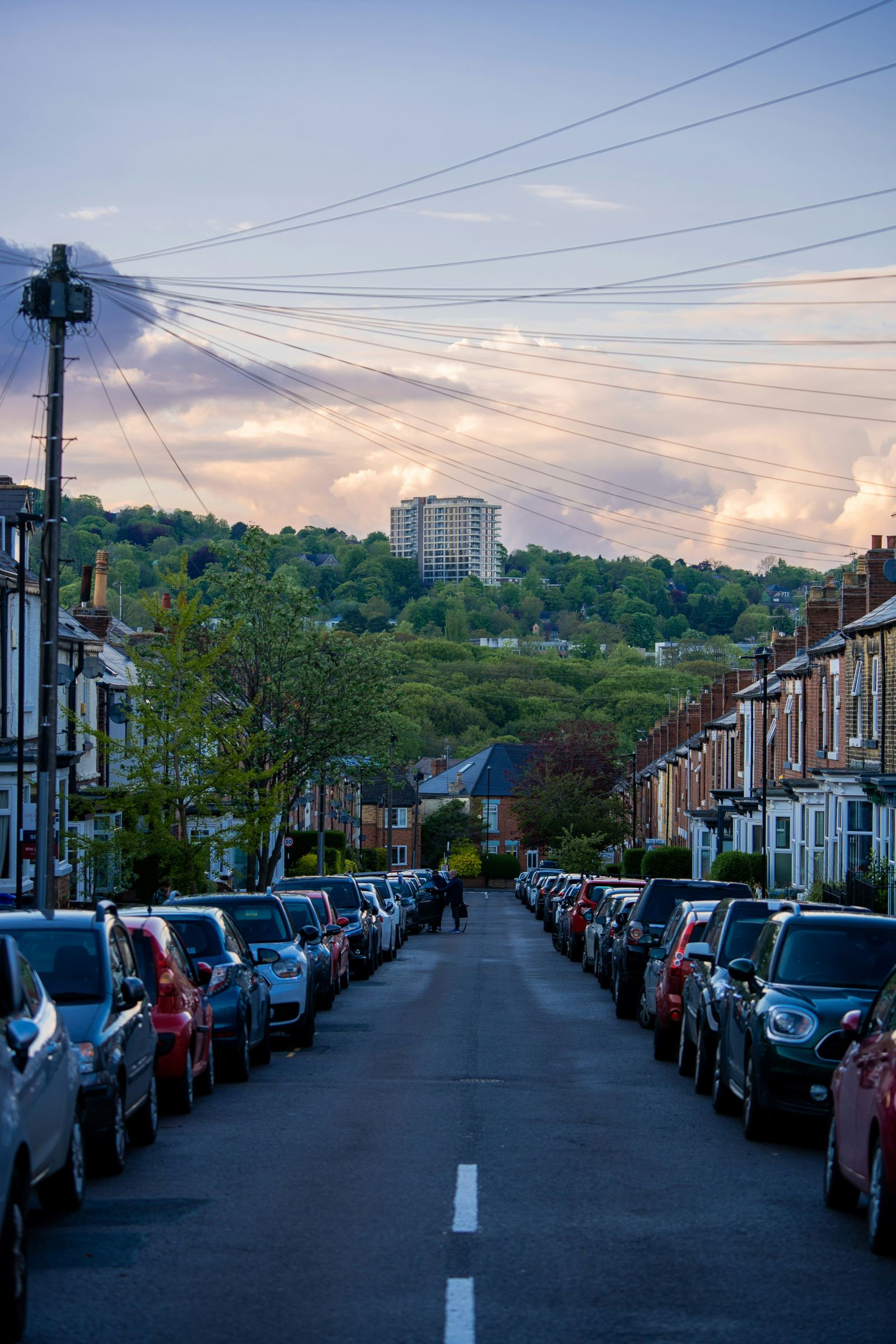 Row of cars parked along a UK residential street at dusk with hills in the background