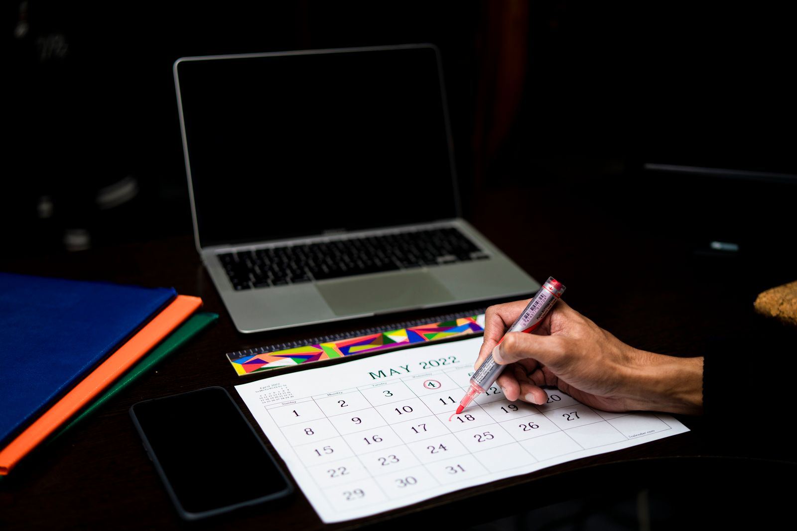 Hand marking a date on a desk calendar beside a laptop and phone